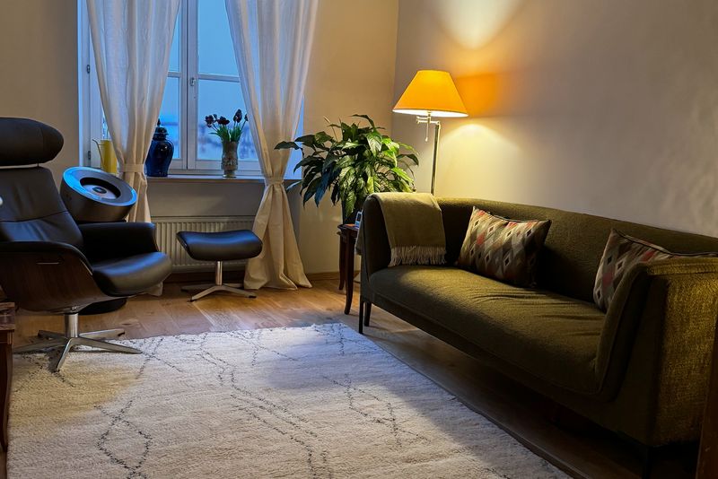 Therapieraum mit grünem Sofa, Ledersessel, hellem Teppich, Stehlampe und Fenster mit Vorhängen